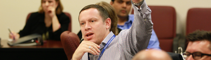 A person in a striped shirt raising their hand in a meeting or classroom setting, with several others seated around in a discussion.