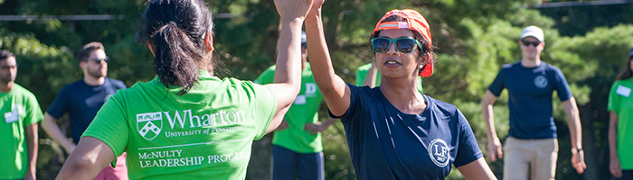 A group of people engaging in an outdoor team-building activity. Two individuals in the foreground are giving each other a high-five, one wearing a green shirt with "Wharton McNulty Leadership Program" written on it, and the other wearing a dark