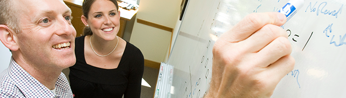 A small group of people engaged in discussion at a whiteboard, with one person writing notes or equations.