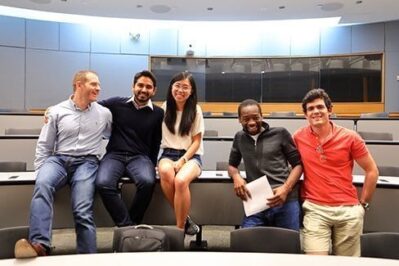 A small group of people sitting on desks in a lecture hall, smiling and posing for the photo. They are casually dressed.