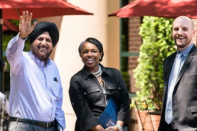 A diverse group of three people stands outside, smiling and engaged. One person waves, and they appear to be in a pleasant, social setting with red umbrellas in the background.