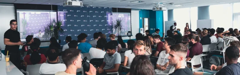 A large group of people seated in a modern conference room, engaged in discussion or listening to a presentation. The room features projection screens and bright lighting.