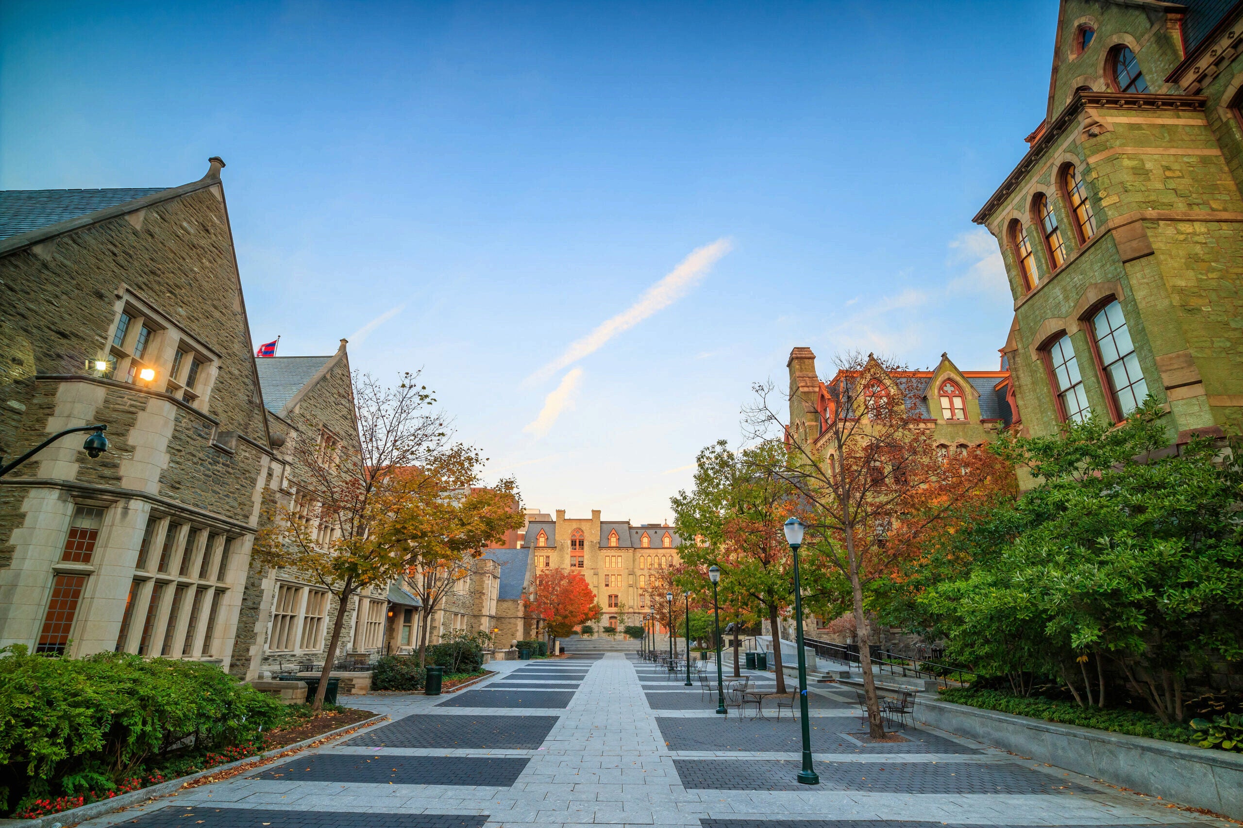 A scenic view of a university campus walkway lined with historic stone buildings featuring gabled roofs. Trees with autumn foliage flank the path.