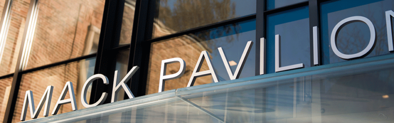 The image displays the exterior of a building with large glass windows and the name "Mack Pavilion" in prominent white letters.
