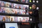 A person stands in front of a large screen displaying multiple video call participants. The Ford logo is visible on a screen below, and a digital clock shows a time.