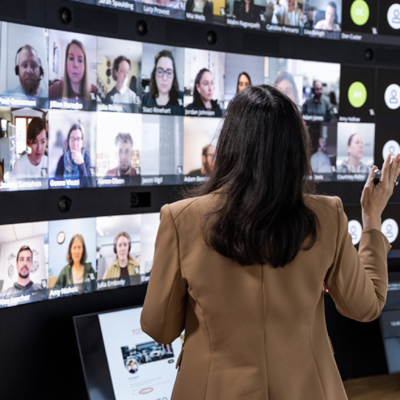Screen Shot 2023 06 22 At 1.23.35 PM A person in a beige blazer is gesturing towards a video wall displaying multiple participants in a virtual meeting.