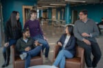 A small group of people gathered in a modern office setting, engaged in conversation and smiling, with tables and chairs in the background.