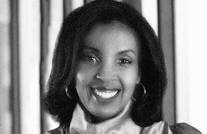 Erika James Black and white headshot of a person smiling, wearing hoop earrings. The background has vertical blurred lines.