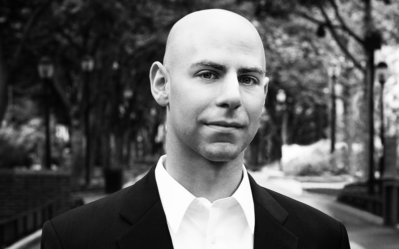 Headshot of a person in a suit, standing outdoors with a blurred background of trees and a pathway. The image is in black and white.