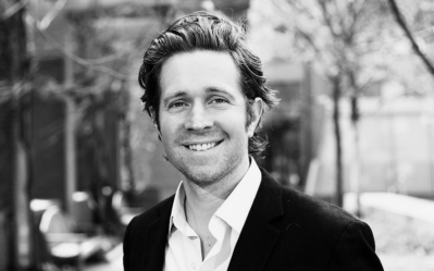 A black and white headshot of a person outdoors, smiling, wearing a suit jacket and a collared shirt, with trees and a building in the background.