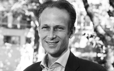 Headshot of a smiling person in a suit, standing outdoors with trees and blurred background. Black and white image.