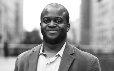 Headshot of a person smiling, wearing a blazer and shirt with an urban cityscape blurred in the background. Black and white photograph.