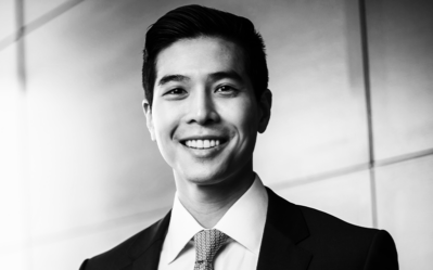 Headshot of a smiling person in a suit and tie, set against a blurred, modern background.