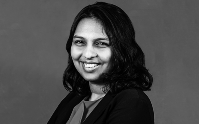 A black and white headshot of a smiling individual with shoulder-length hair, wearing a dark blazer and a top.