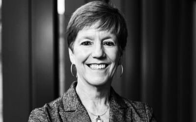 A black and white headshot of a smiling person with short hair, wearing a blazer and hoop earrings.