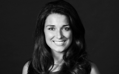 A black and white headshot of a person with long, wavy hair and a bright smile, set against a dark background.