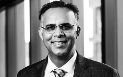 Headshot of a smiling person in a suit and tie, wearing glasses, with a blurred indoor background.