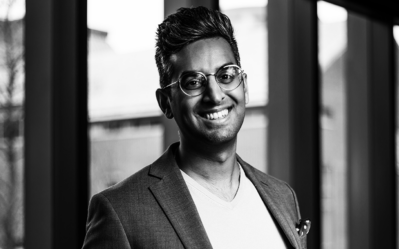 A person wearing glasses and a blazer, smiling, and standing indoors in front of large windows. The image is in black and white.