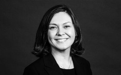 Headshot of a person smiling, with short hair and wearing a dark blazer, set against a plain background.