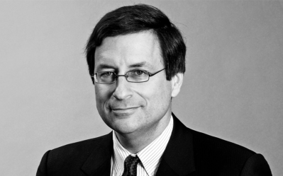 Headshot of a person with glasses, wearing a suit and tie, smiling slightly against a neutral background.