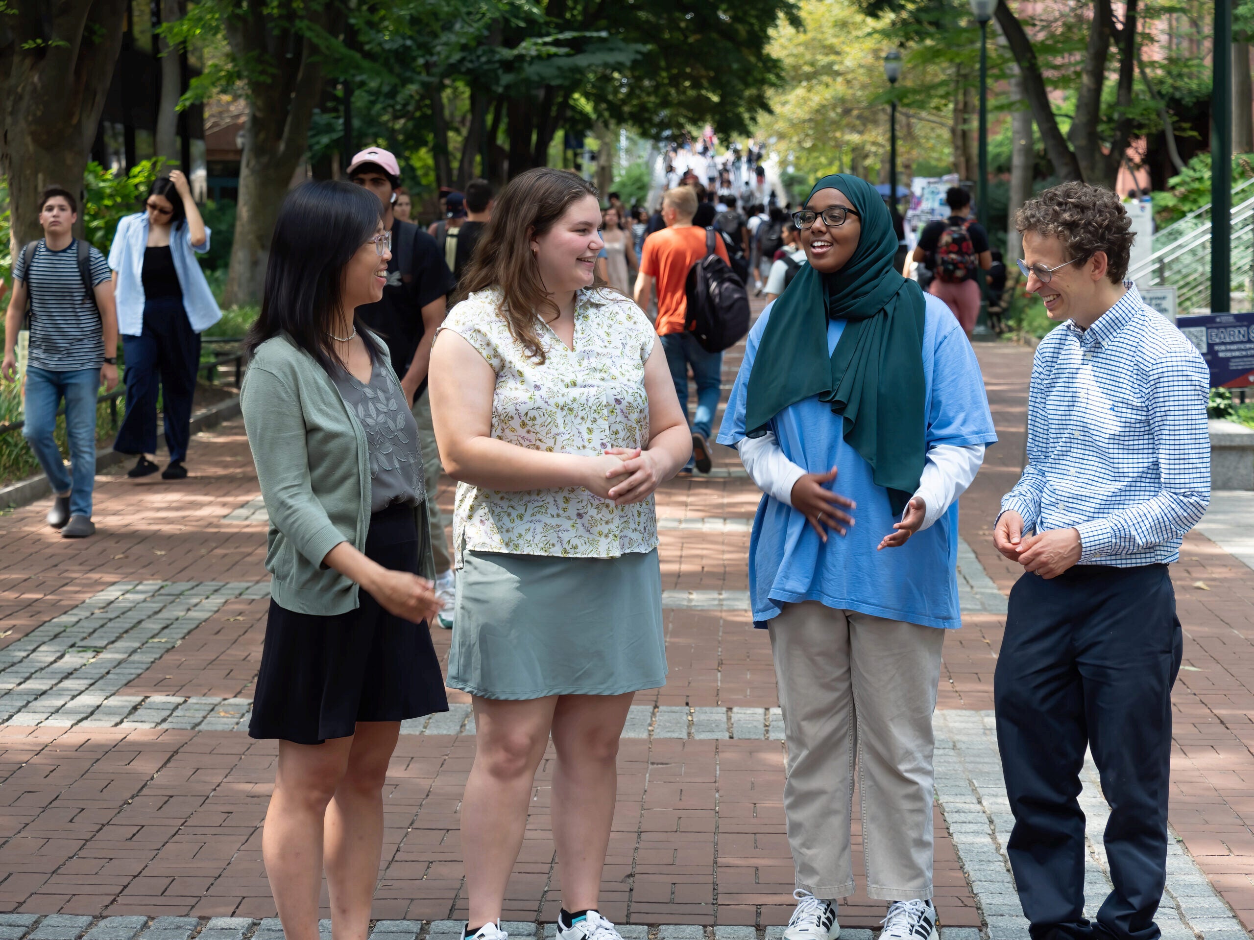 P9069676 A small group of people stand together outdoors, smiling and engaging in conversation. The setting is a brick pathway lined with trees, creating a campus-like atmosphere.