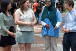 A diverse group of people standing and talking on a tree-lined pedestrian street or pathway, with more individuals in the background.