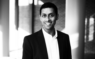 Black and white headshot of a person in a suit, smiling, with a blurred background of windows and walls.