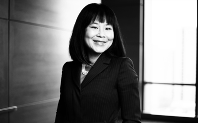 Headshot of a person smiling, wearing a suit and pearls, with a blurred indoor background and natural light coming from a window.