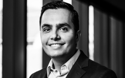 Black and white headshot of a person wearing a suit, smiling, and standing in front of a blurred background with vertical elements.