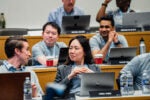 A group of people attentively participating in a lecture or seminar, seated at desks with laptops and bottled water. They appear engaged in conversation or discussion.
