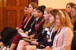 A group of people sitting in a row at an event or conference, focused and taking notes, dressed in professional attire.
