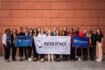 A group of people is standing together, holding banners that read "Resilience Education" and "Wharton University of Pennsylvania," in front of a brick wall.