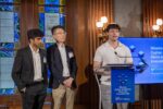 Three individuals are presenting at the Wharton Cypher Accelerator Demo and Innovation Day. The setting includes a podium, a blue event banner, and stained glass windows in the background.