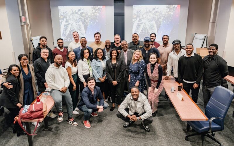 A diverse group of people gathered in a conference room, smiling and posing for a group photo. Two screens in the background display congratulatory messages.
