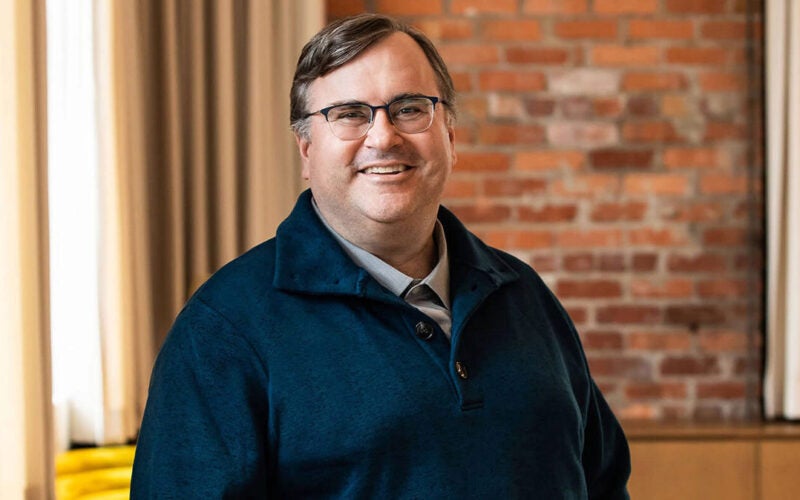 A person wearing glasses and a dark blue pullover stands smiling in an interior setting with beige curtains and a brick wall.