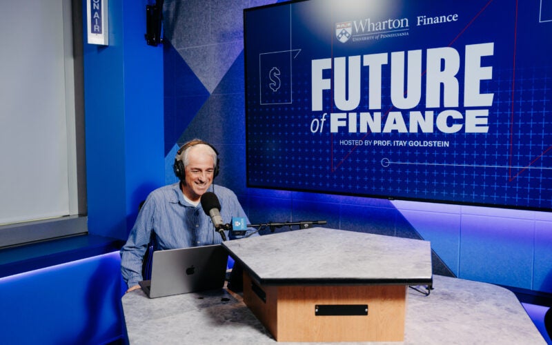 A person wearing headphones is speaking into a microphone in a recording studio. They are seated at a desk with a laptop. Behind them is a large screen displaying the title "Future of Finance," indicating a podcast or radio show setting.