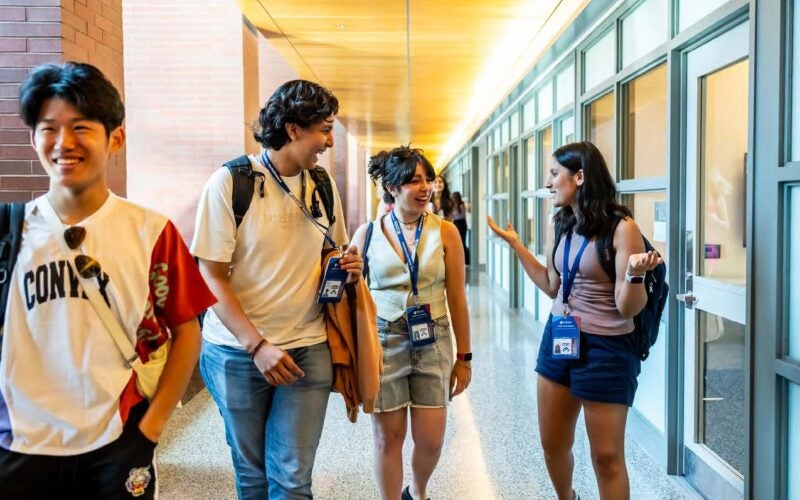 A group of young people walking and talking in a bright hallway, wearing casual clothing and identification lanyards.