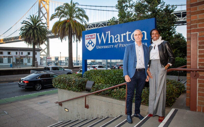 A couple of people standing in front of a Wharton University of Pennsylvania sign, with a bridge and palm trees in the background.