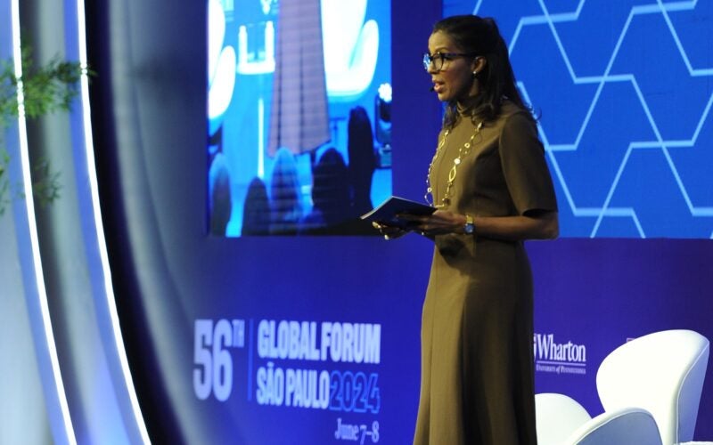 A person in a brown dress is speaking on stage at the 56th Global Forum in São Paulo 2024. The backdrop is blue with geometric patterns and a large screen.