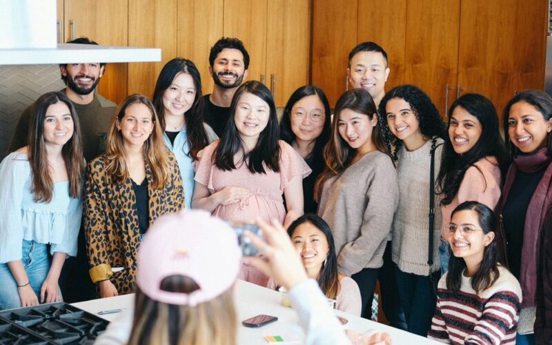A group of people smiling around a kitchen counter, with one person in the foreground taking a photo. They appear to be gathered for a celebration or casual event.