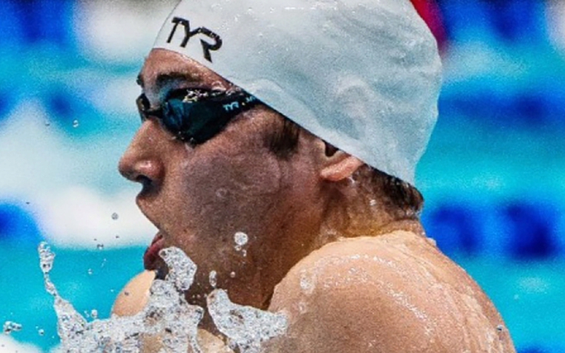 A swimmer wearing a white swim cap and dark goggles is emerging from the water, focusing intensely. The background shows a swimming pool with blurred lane markings.