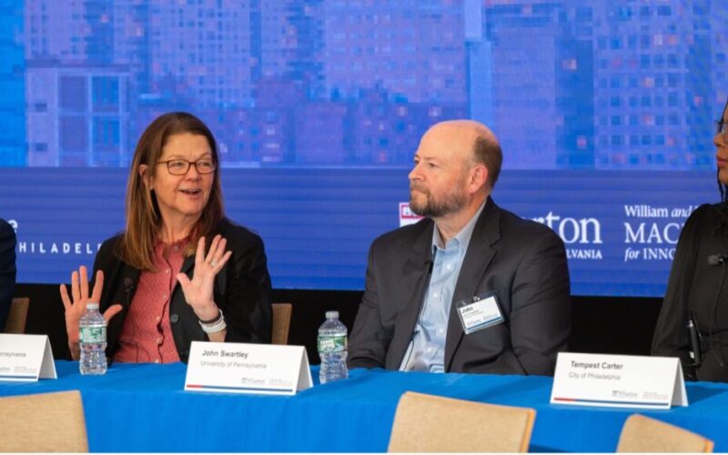 A panel discussion with four individuals sitting at a table with microphones and name cards. They appear engaged in conversation against a backdrop of a cityscape.