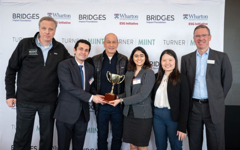 A group of people stands together, smiling, while holding a trophy. They are in front of a backdrop with logos for the Bridges Impact Foundation, Wharton, and Turner MIINT, indicating a likely celebratory or award-winning event.