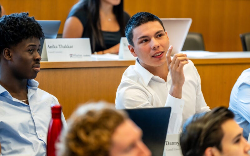 A group of students in a classroom setting, some taking notes and others engaging in discussion. Nameplates and laptops are visible.