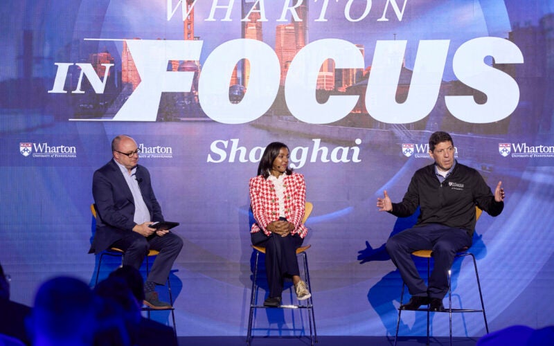 A panel discussion is taking place with three individuals seated on stage. The backdrop displays the words "Wharton in Focus Shanghai" with Wharton University branding.