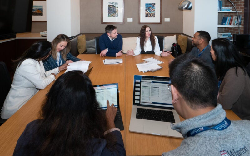A group of people having a discussion in a conference room. They are seated around a wooden table with laptops and documents. The room has framed artwork on the walls and shelves with books.