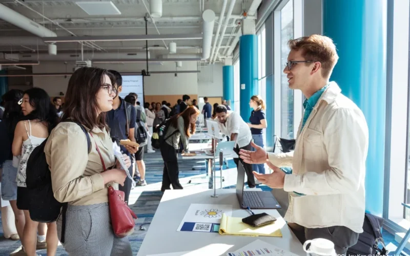 A small group of people engaging in conversation at a professional networking event or job fair. Tables with laptops and informational materials are visible, suggesting a business or educational setting.