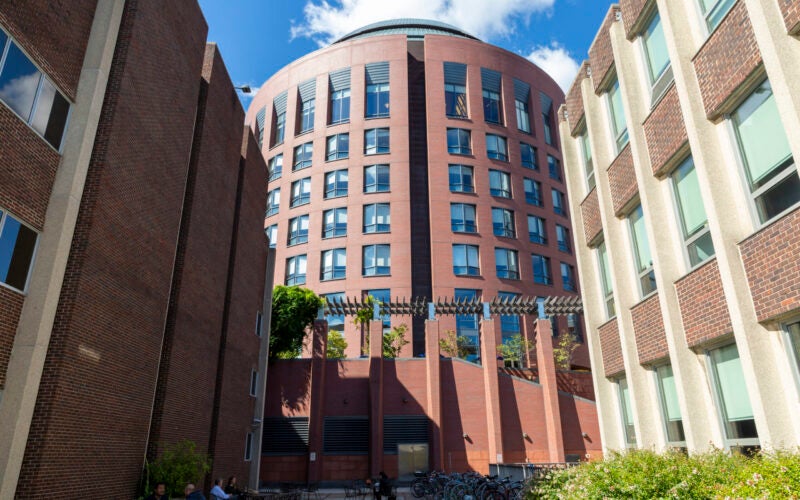 A modern, cylindrical brick building surrounded by other brick structures. The scene includes outdoor seating and bicycles, under a clear blue sky.