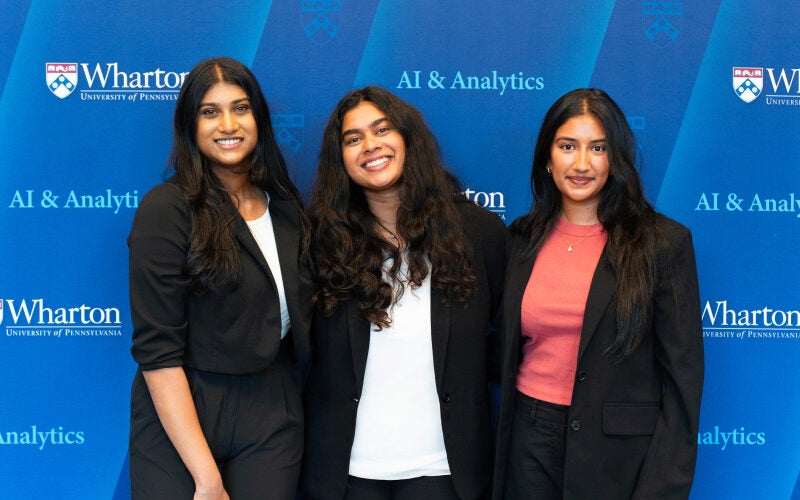 Three individuals standing in front of a blue backdrop with "Wharton University of Pennsylvania AI & Analytics" logos. They are dressed in business attire.