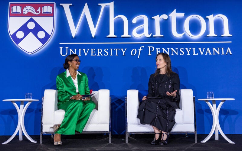 Two individuals seated on stage at a Wharton University of Pennsylvania event. The backdrop displays the university's logo.